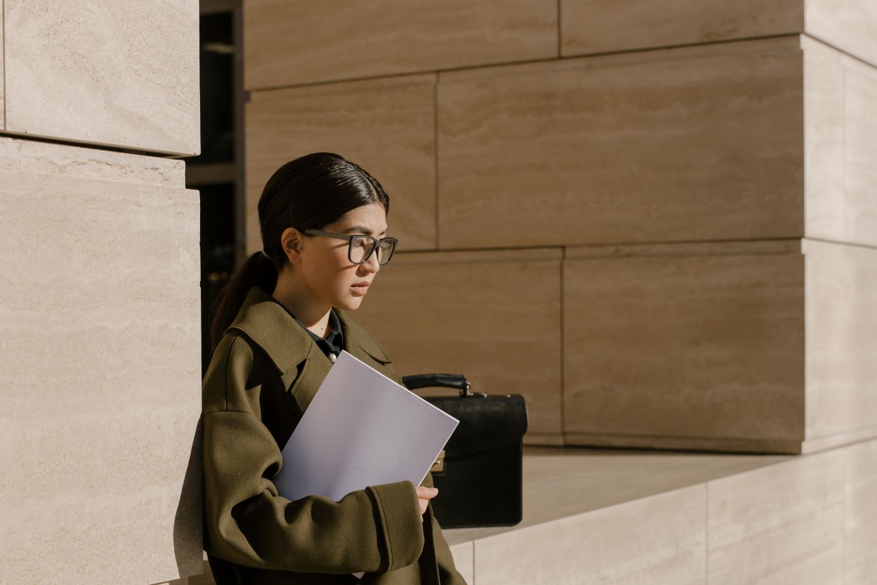 An Asian woman in eyeglasses holding a folder in an urban outdoor setting, exuding professionalism.